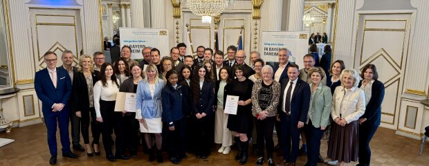 Gruppenfoto im festlich geschmückten Spiegelsaal der Regierung der Oberpfalz. Rund 30 Personen – darunter die Preisträgerinnen und Preisträger des Integrationspreises 2025, Ehrengäste sowie Vertreterinnen und Vertreter der Regierung der Oberpfalz – stehen zusammen, einige halten Urkunden in den Händen. Im Hintergrund sind Banner mit der Aufschrift „Integration leben – In Bayern daheim“ zu sehen.