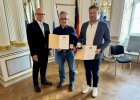 Regierungspräsident Walter Jonas steht neben den Geehrten Stefan Fröhlich und Johannes Meckl, die Urkunden und Medaillen für ihre Lebensrettung in Empfang nehmen.