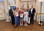 Zwei Schülerinnen des Albrecht-Altdorfer-Gymnasiums halten ihre Urkunden und stehen gemeinsam mit Lehrkräften und Ehrengästen vor den Fahnen.