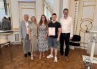 Ein Schüler hält seine Urkunde und steht gemeinsam mit Vertreterinnen und Vertretern von Regierung und Schule im Spiegelsaal.