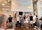 Schülerinnen und Schüler stehen mit einer Moderatorin vor Publikum auf der Bühne im Spiegelsaal.