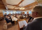 Blick über die Schulter des Redners in einen voll besetzten Festsaal mit langen Tischreihen.
