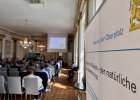Ein Informationsaufsteller der Regierung der Oberpfalz zum Thema Boden, Gestein und Grundwasserschutz steht im Vordergrund. Im Hintergrund sitzen Teilnehmerinnen und Teilnehmer im Spiegelsaal und verfolgen einen Vortrag.